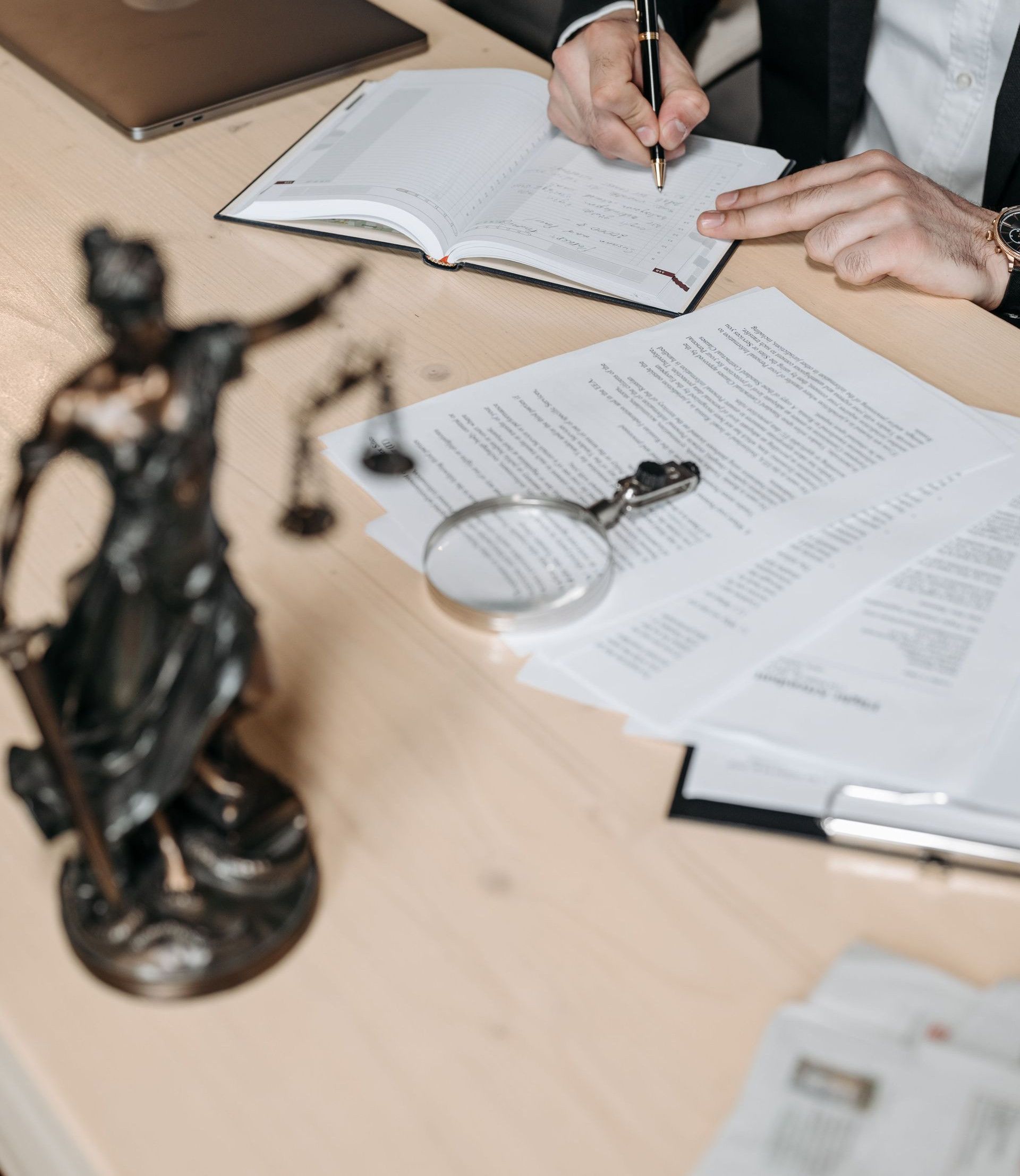 Professional working on documents at desk with legal statue and magnifying glass.